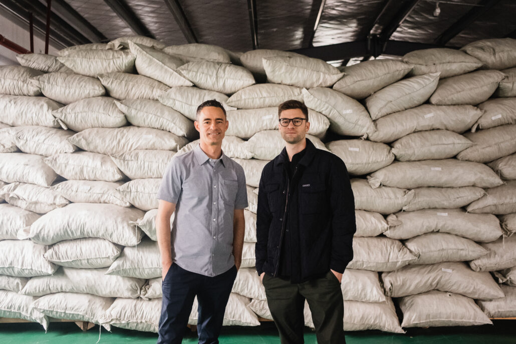 Two men standing in front of a pile of sacks.